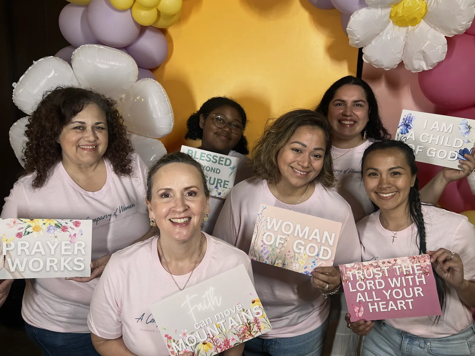 Smiling women in pink A Company of Women shirts holding faith signs in front of a balloon flower arch at the Spring market